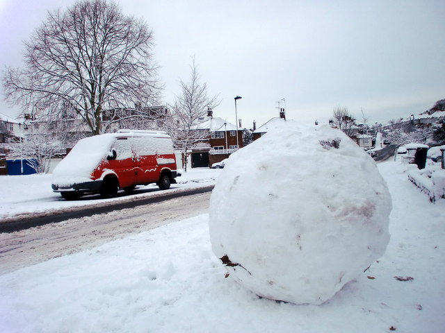 A big snowball the size of a boulder with a red van next to it on the street