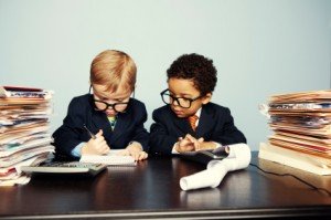 Two young kids dressed in fancy suits pretending to be accountants wearing glasses with stacks of papers and folders on the desk. 